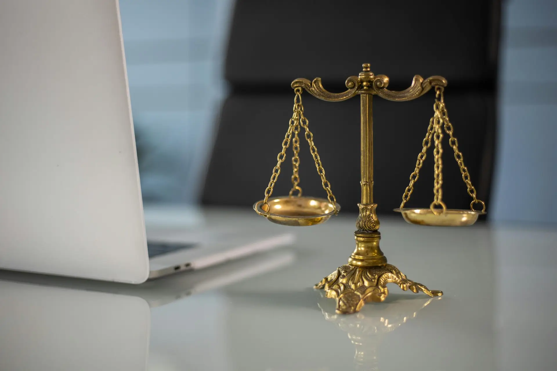 Balance dorée de la justice posée sur un bureau en verre, à côté d’un ordinateur portable.
